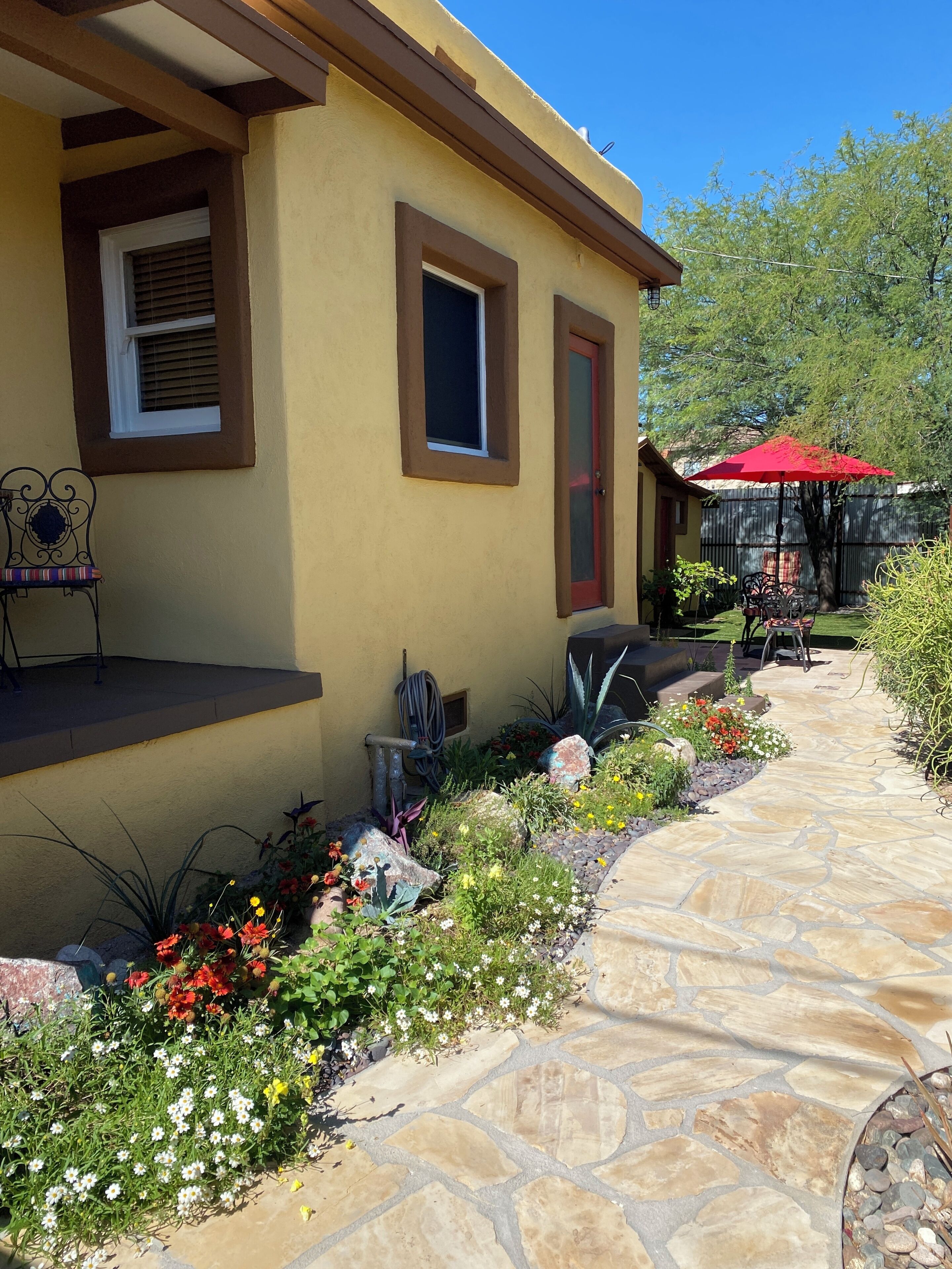 A yellow building with a stone pathway, a red umbrella, and various plants.