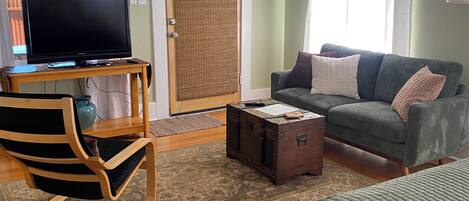 Une pièce avec un téléviseur posé sur un meuble en bois, un canapé vert avec des coussins, un tapis à motifs et une commode en bois.