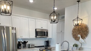 A modern kitchen with white cabinets, stainless steel appliances, a central island with bar stools, and decorative wall art. - Sweeping Vue of the Gulf of Mexico, 30 steps to beach (Mexico Beach)