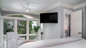 Ein Schlafzimmer mit einem großen Bett, einem flachbildfernseher an der Wand und Blick ins Freie durch ein großes Fenster.