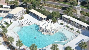 Eine Vogelperspektive zeigt ein Resort mit mehreren Schwimmbecken, Liegestühlen und überdachten Sitzbereichen.