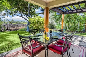 Un espace de restauration en plein air avec une table dressée pour quatre personnes, entourée de chaises dotées de coussins rouges, sous une pergola offrant une vue sur un jardin luxuriant et un paysage lointain.