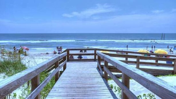 A wooden boardwalk leading to a beach with umbrellas and people.