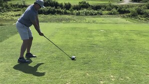 Ein Golfplatz mit einer Person, die bereit ist, einen Golfball zu schlagen, umgeben von üppiger Vegetation und Bergen im Hintergrund.