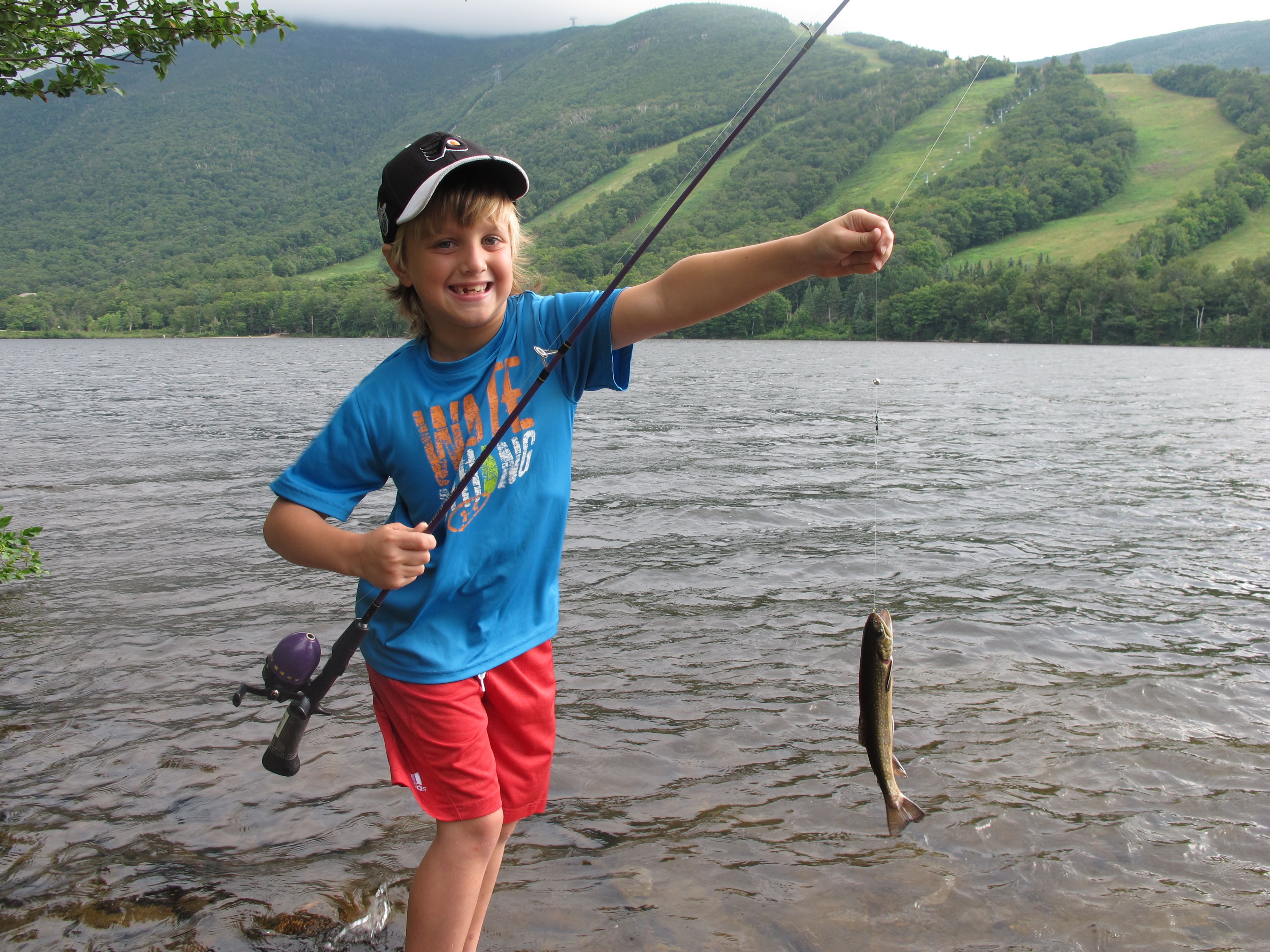 A person holding a fishing rod with a fish on the line, standing by a lake with hills in the background.