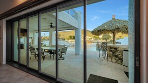 Una vivienda moderna con un patio de cristal que incluye zona de comedor, piscina y una construcción con techo de paja.