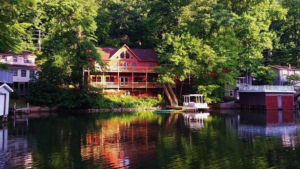 A two-story house with a red roof and a balcony, situated by a calm lake with a dock and a small boat.