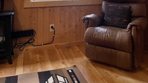 A room with wooden walls, a large window with a view of trees, a leather armchair with a patterned cushion, a flat-screen TV, and a power outlet on the floor.