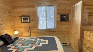A cozy wooden cabin bedroom with a bed, a nightstand, a dresser, and a window with blinds.