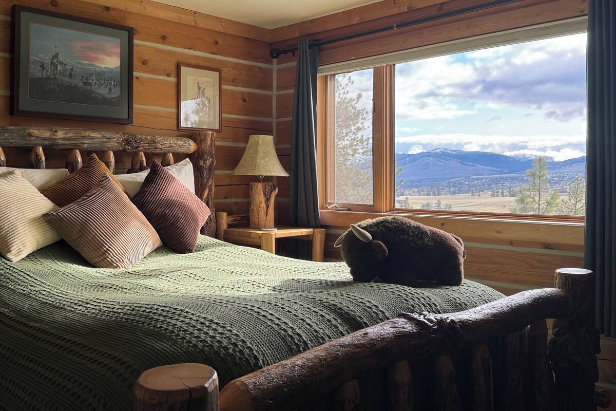 A cozy log cabin bedroom with a large bed, wooden headboard, and a view of mountains through a window.