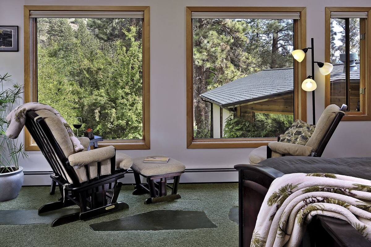 A room with a rocking chair, a small table, and a bed with a blanket. There are large windows with a view of trees and a building outside.