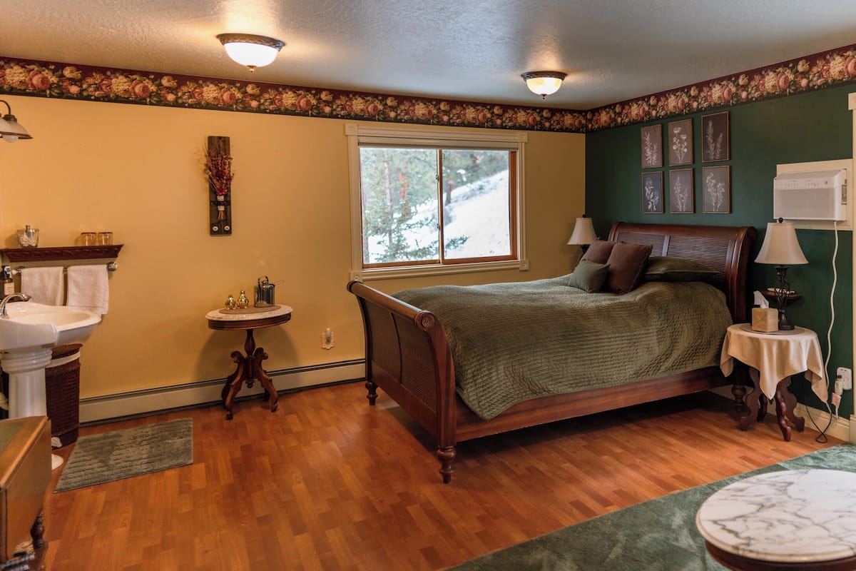 A bedroom with a wooden bed, a window with a view of trees, a sink, and a small table.