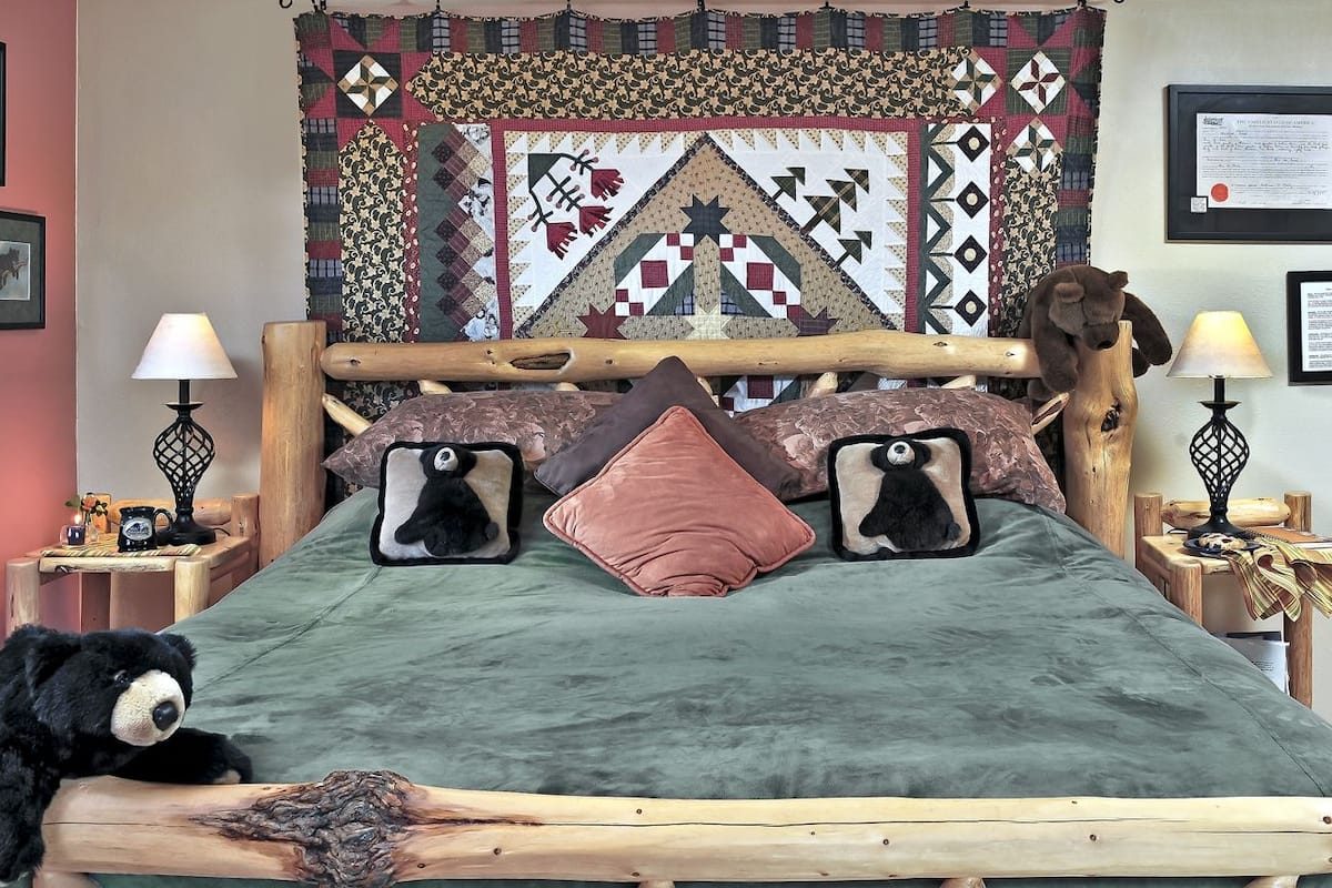 A bed with a wooden headboard, two black and white pillows, a green bedspread, and a quilt hanging on the wall behind.