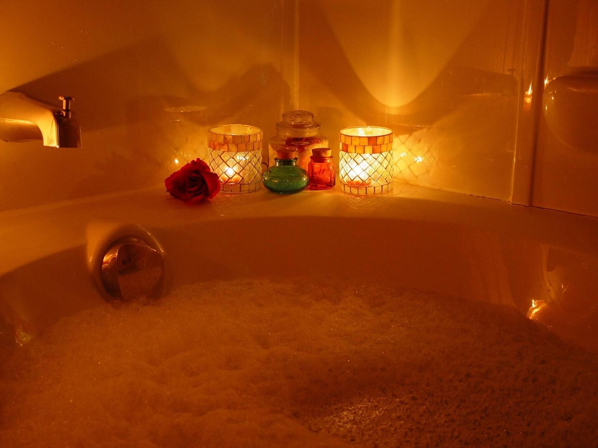 A bathtub filled with bubbles, surrounded by three lit candles and a red rose.