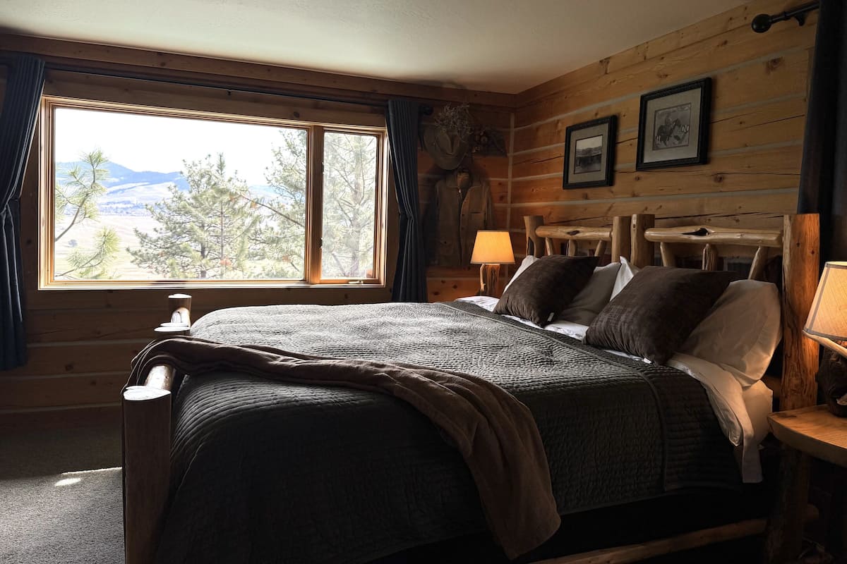 A wooden cabin-style room with a large bed, two bedside lamps, a window with a view of trees and mountains, and a framed picture on the wall.