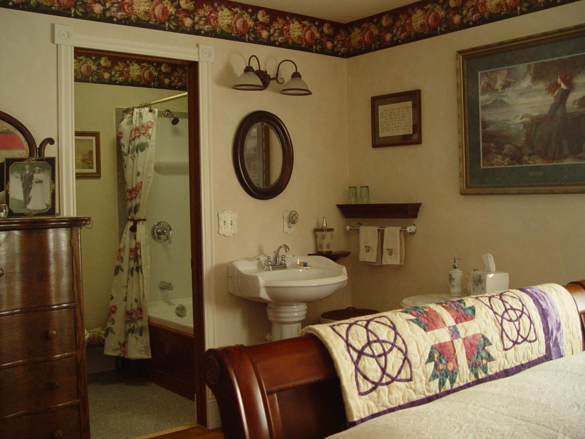 A bedroom with a wooden dresser, a white sink, a mirror, a bed with a quilt, and a painting on the wall.