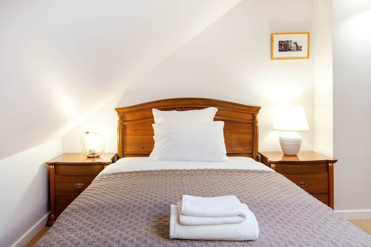Una cama bien hecha con cabecera de madera, una lámpara de noche y una toalla doblada sobre la mesita.