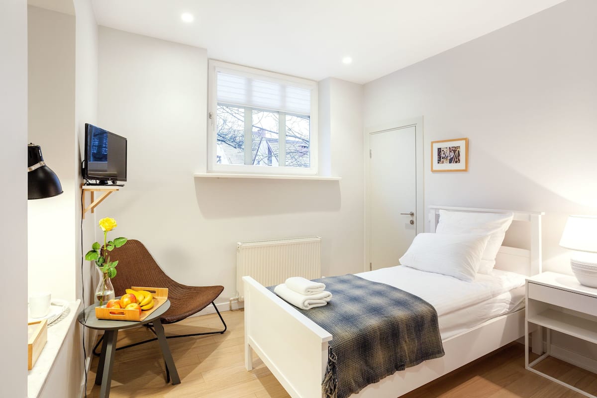 Un dormitorio moderno con una cama, una silla, una mesita con fruta, un radiador, una ventana y un cuadro en la pared.