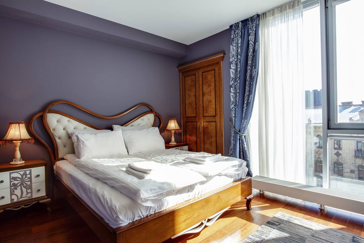 Una cama bien hecha en una habitación con paredes moradas, un armario de madera y un ventanal con cortinas.