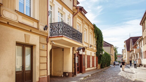 A cobblestone street lined with historic buildings, featuring balconies with wrought iron railings and a mix of architectural styles. - Jolly apartments (Vilnius)