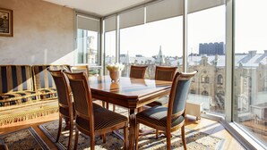 A dining area with a wooden table and chairs, a vase with flowers, and a view of buildings through large windows. - Jolly apartments (Vilnius)