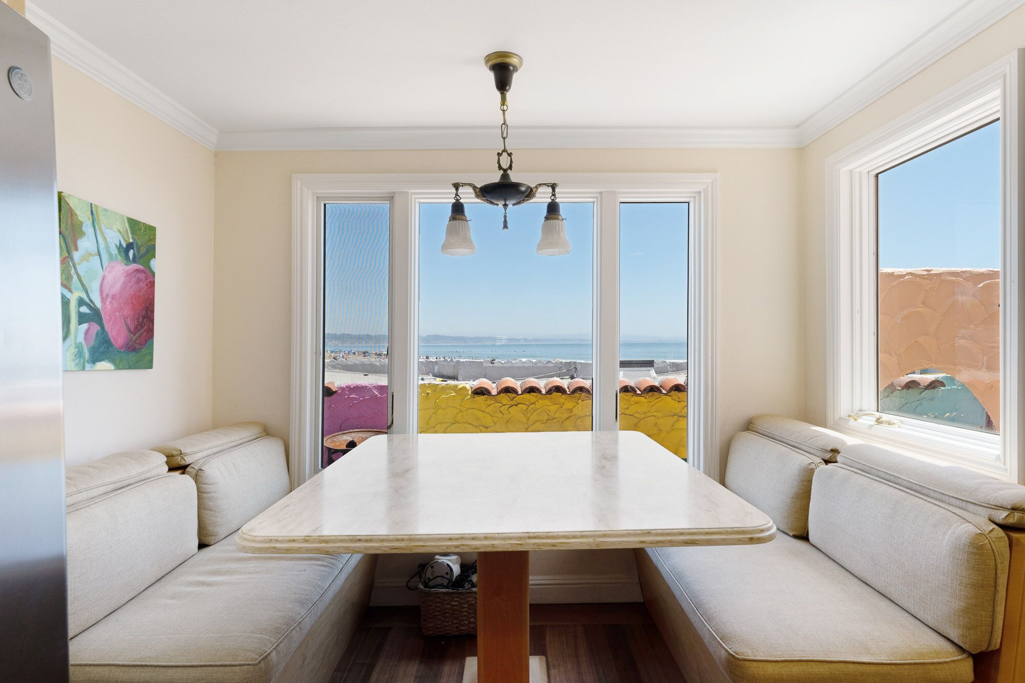 A dining area with a table and benches, large windows with a view of the beach, and a ceiling light fixture.