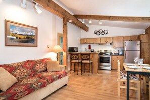 Een gezellige woonkamer met een gestreept bankje, een eettafel met stoelen, een keuken met houten kastjes en apparatuur, en een lijst met een foto aan de muur.