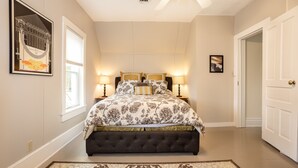 Ein Schlafzimmer mit einem großen Bett, zwei Nachttischlampen, einem gerahmten Bild an der Wand und einer Tür rechts.