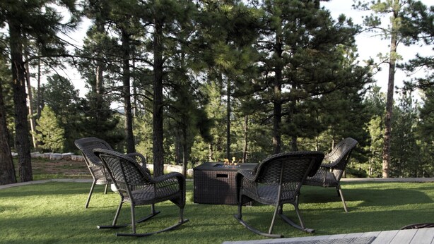 An outdoor seating area with wicker chairs and a table, surrounded by a grassy lawn and tall trees.