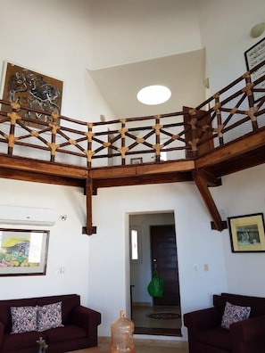 Un vaste espace de vie doté d’un haut plafond et d’un balcon en bois. On y trouve deux canapés avec des coussins à motifs, un grand distributeur d’eau et des œuvres d’art accrochées aux murs.