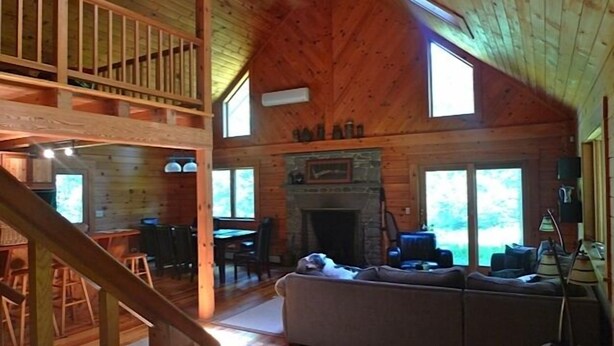 L’intérieur d’un chalet en bois comprend une chambre à coucher située en mezzanine, un salon avec un canapé, une cheminée et de grandes fenêtres.