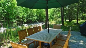 Une table et des chaises en bois sont disposées à l’extérieur, sous un parasol vert, et sont entourées d’une végétation luxuriante et d’un petit ruisseau.