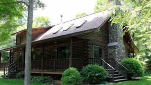 Une cabane en rondins avec une cheminée en pierre, une véranda et un escalier menant à l’entrée.