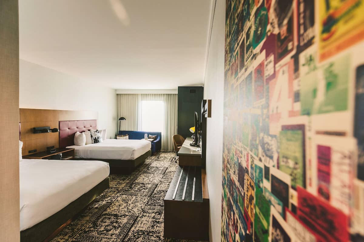 Ein Hotelzimmer mit zwei Betten, einem Schreibtisch, einem Stuhl, einem Fernseher und einem großen Poster an der Wand.