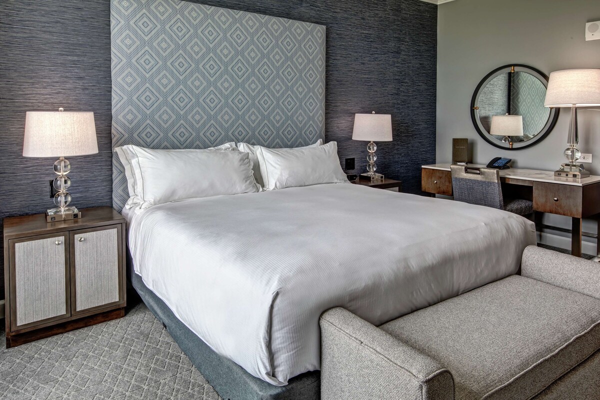 Una habitación de hotel moderna con una cama grande, dos mesitas de noche con lámparas, un escritorio con una silla y un espejo redondo.