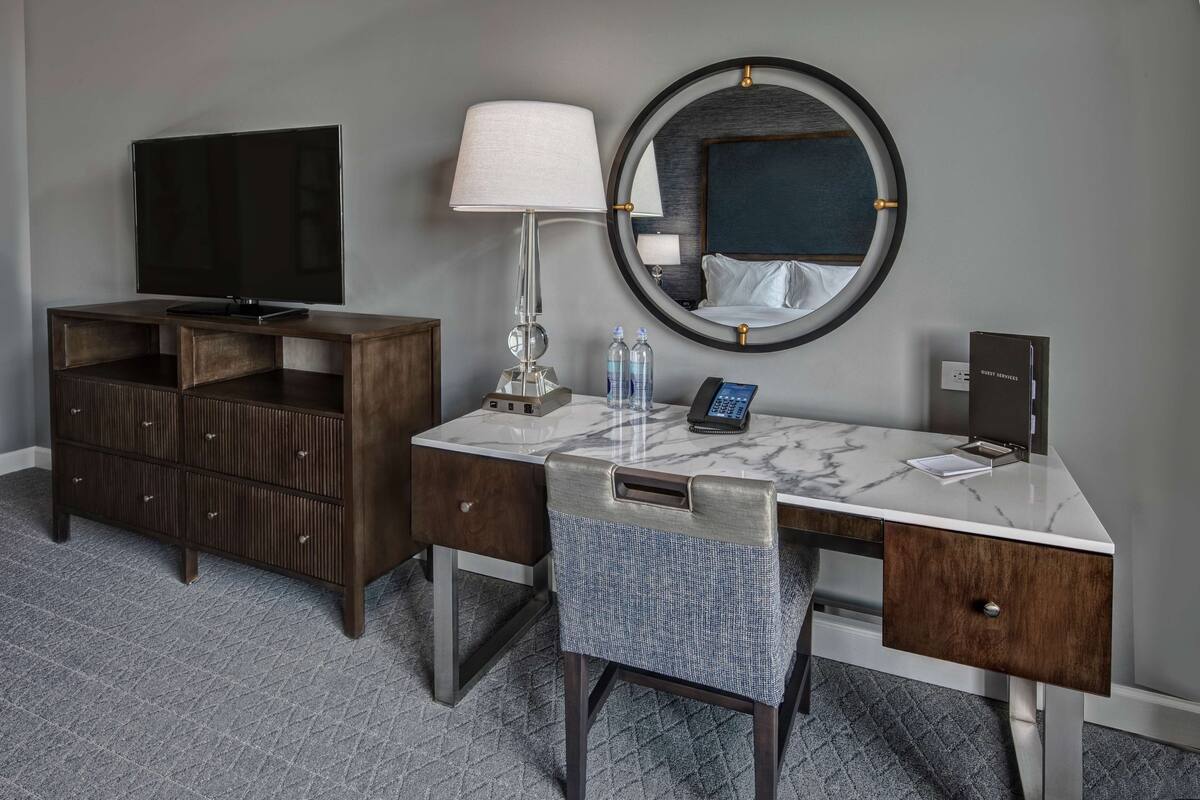 Habitación de hotel con un escritorio de mármol, televisor, lámpara, espejo y una silla.
