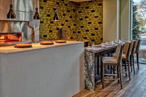 Moderni keittiö, jossa on pizzauuni, pizzatarjoilutisk sekä ruokailutila tuoleineen ja pöytineen.