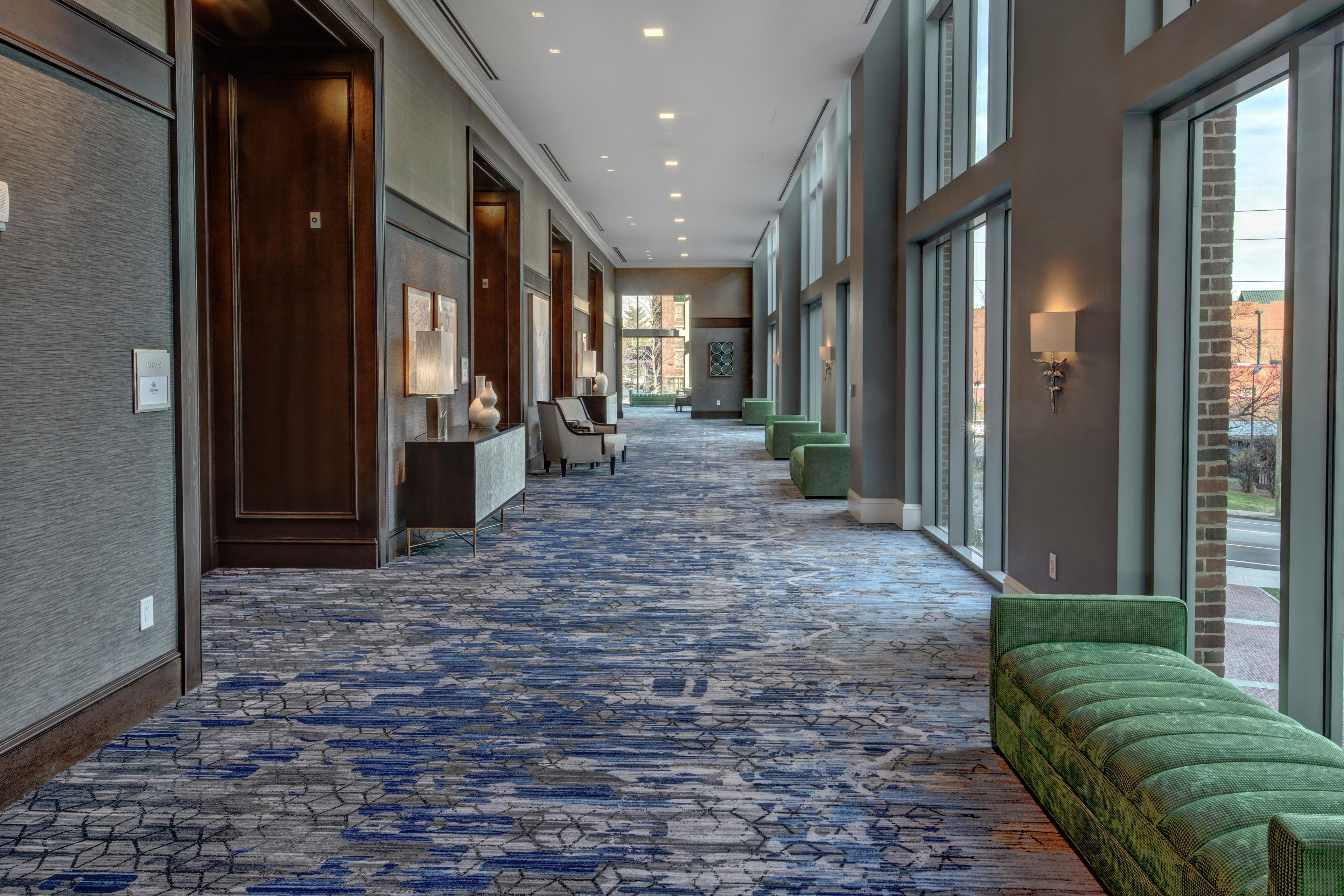 Un pasillo de hotel con alfombra estampada, zonas de estar y amplios ventanales.