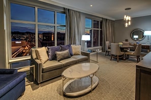 Ein Hotelzimmer mit großem Fenster, einer Couch, einem Couchtisch, einem Essbereich mit Stühlen und einer Lampe.