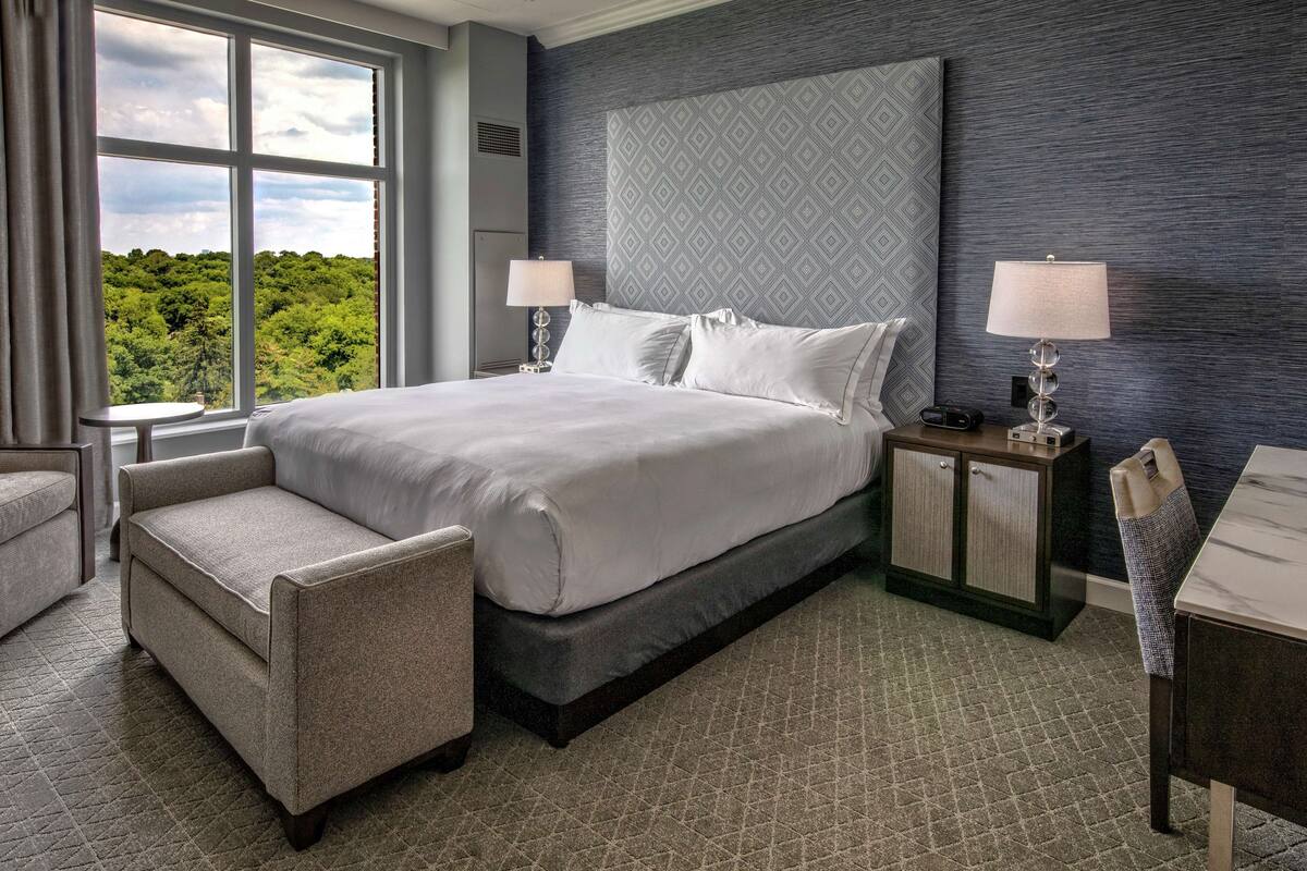 Habitación de hotel con una cama grande, dos mesitas de noche con lámparas, una silla y vistas al exterior.