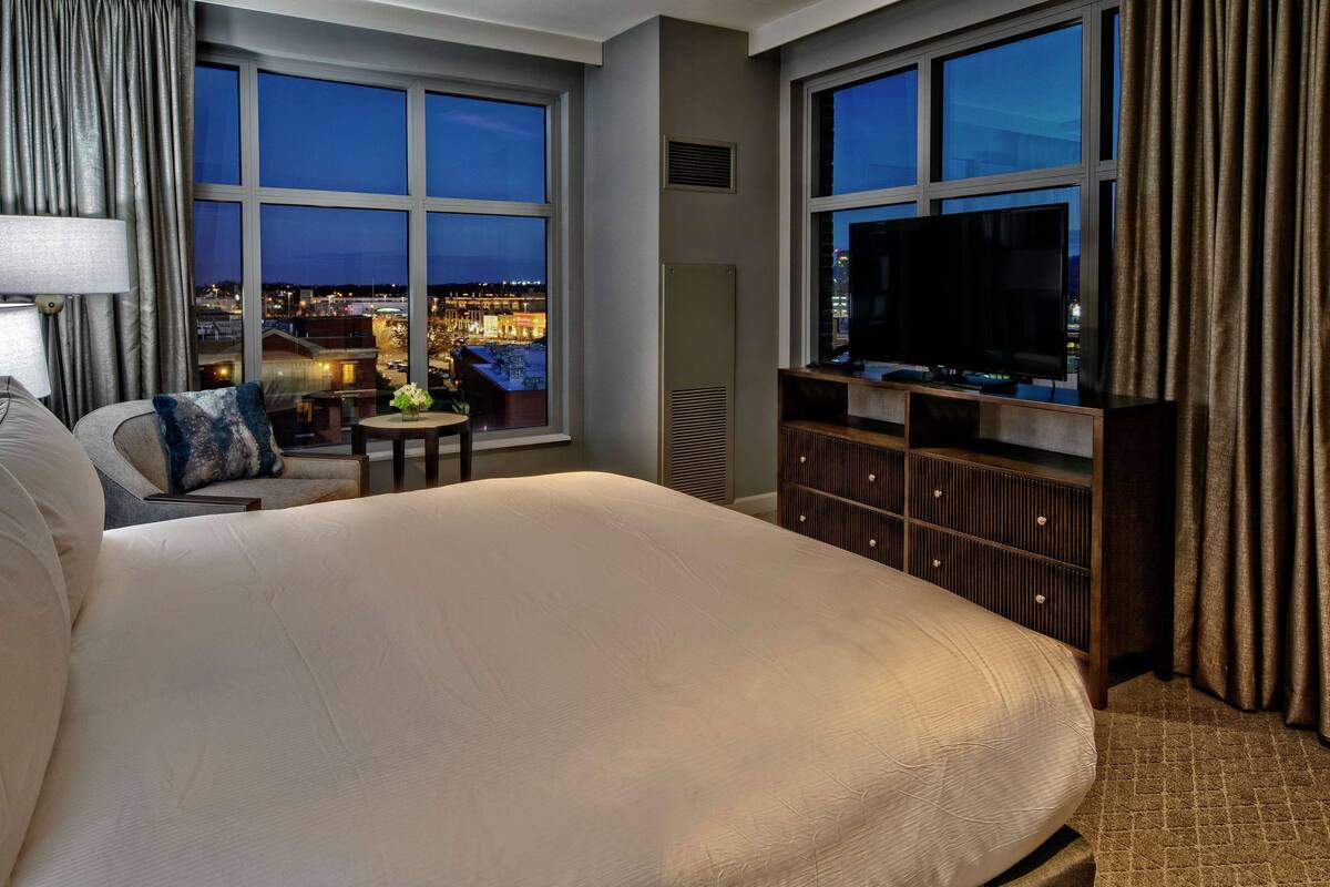 Habitación de hotel con una cama grande, un televisor, una mesita con un florero y vistas a la ciudad al anochecer.