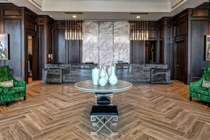 Een hotel lobby met een marmeren receptiebalie, twee groene fluweel fauteuils, een centrale tafel met vazen, en een visgraat parketvloer.