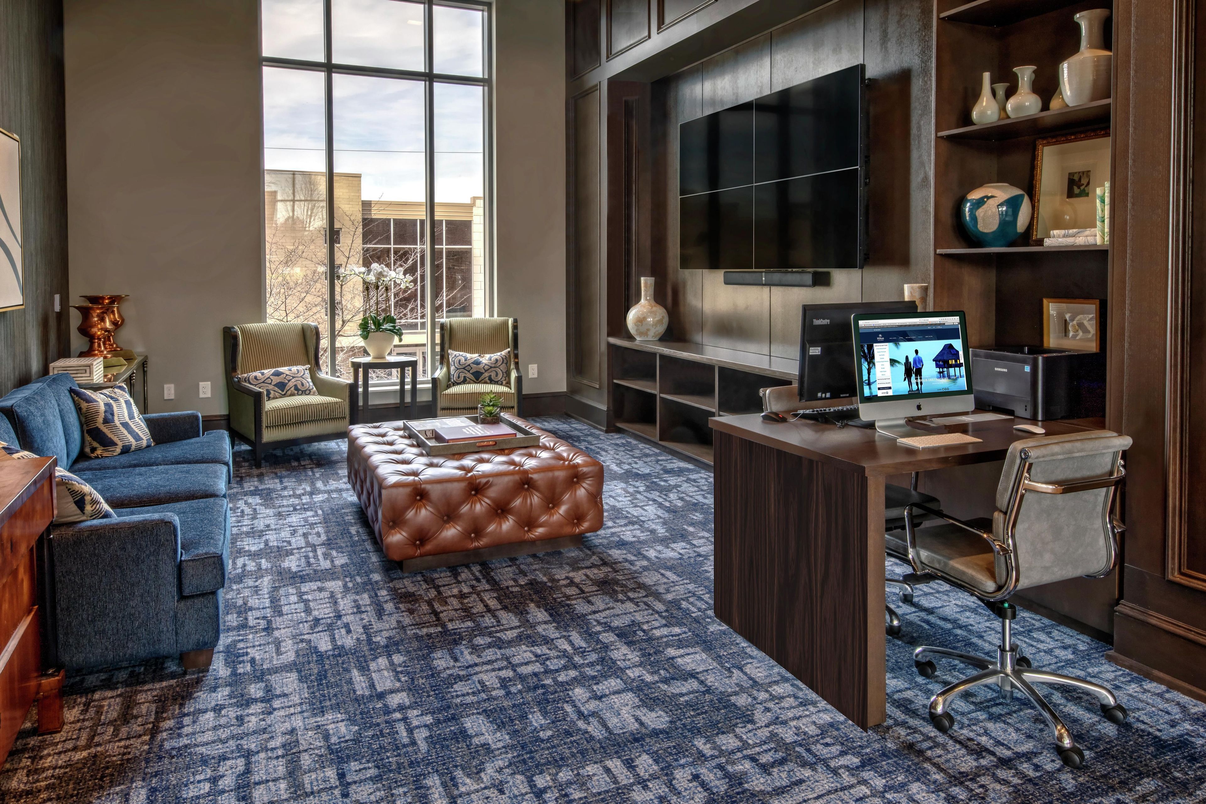 A spacious living room with a large window, a flat-screen TV, a desk with a computer, and a comfortable seating area with a sofa and ottoman.