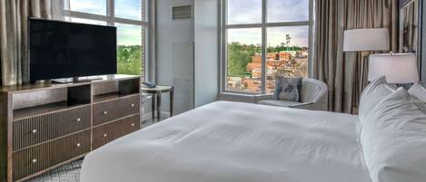 A hotel room with a large bed, a flat-screen TV on a wooden console, a desk with a chair, and a view of the city through large windows.