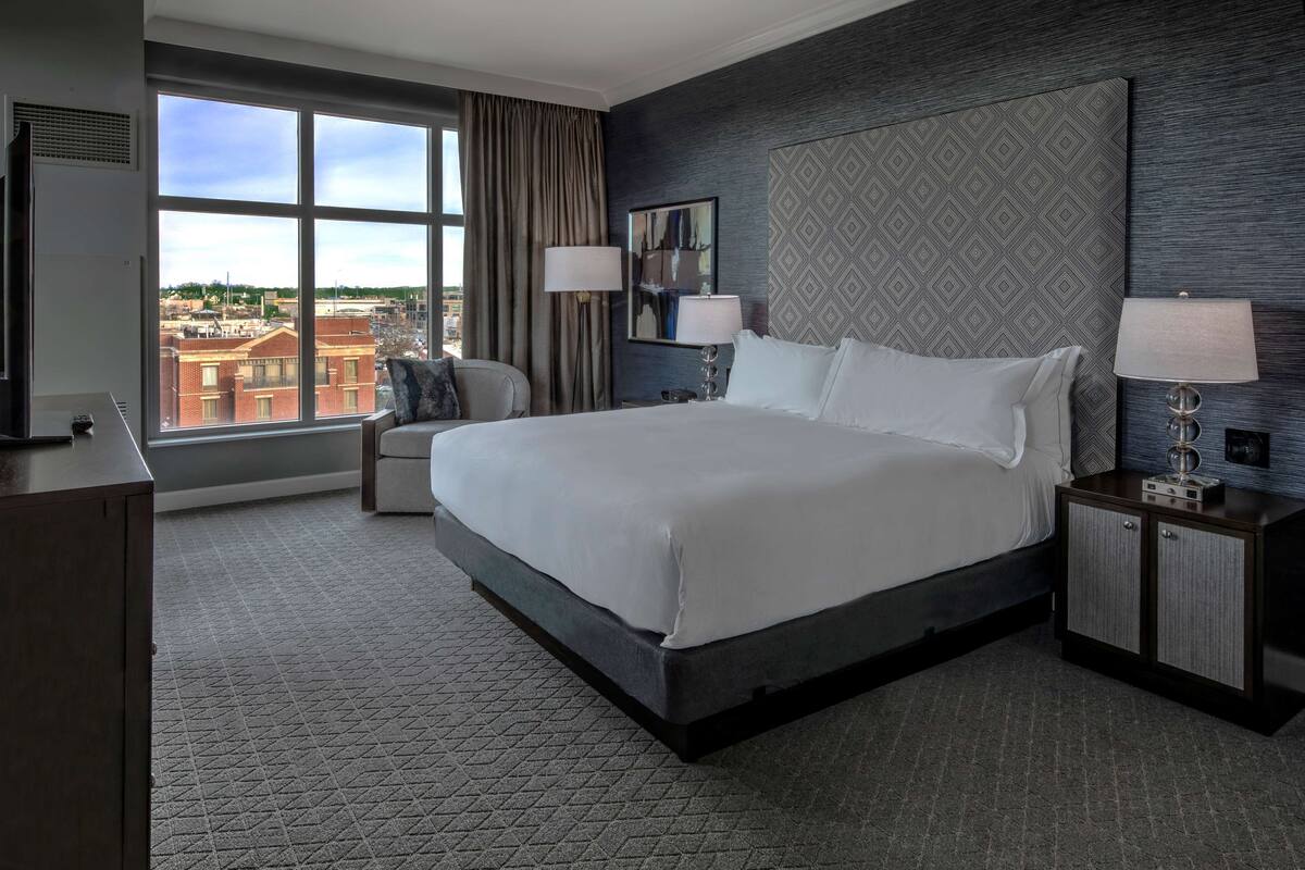 Un cuarto de hotel con una cama grande, un escritorio, una silla, una mesita de noche con lámpara, y vistas de la ciudad a través de un ventanal amplio.