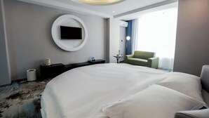 Een moderne hotelkamer met een groot bed, een rond aan de muur bevestigd televisietoestel, een klein bijzettafeltje, een groene fauteuil en uitzicht buiten door een raam.