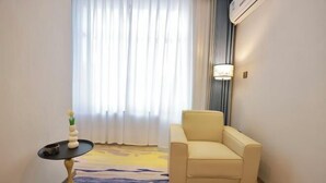 Een hotelkamer met een beige fauteuil, een klein rond tafeltje met een vaas en een groot raam met dunne gordijnen.