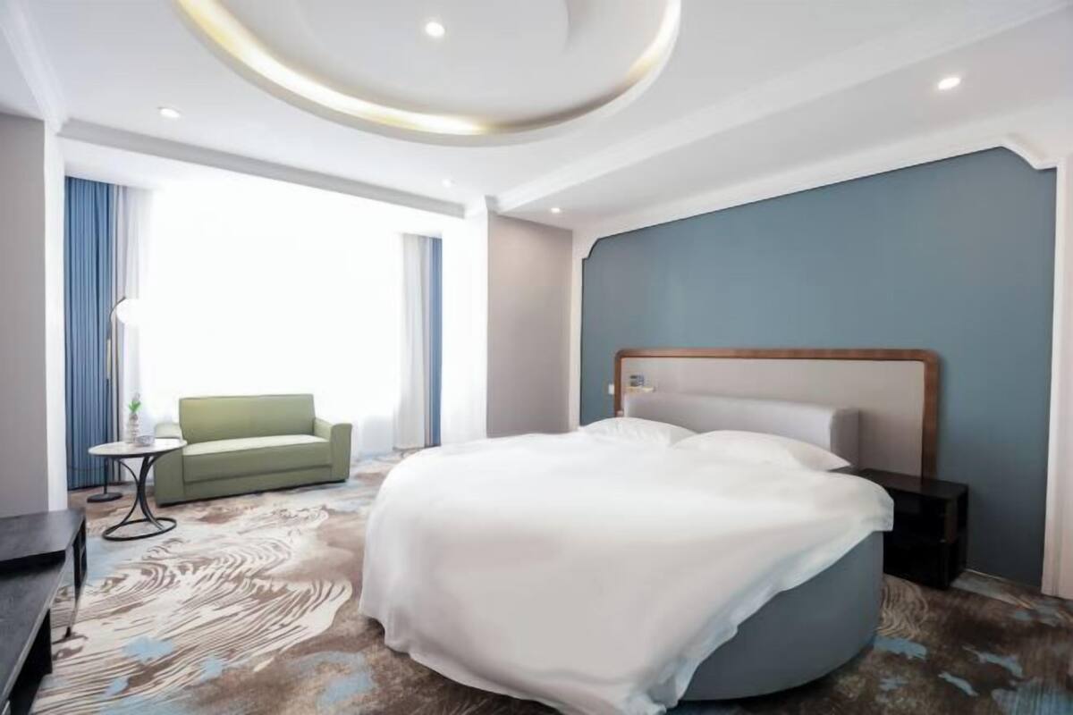 Moderni hotellihuone, jossa on pyöreä sänky, vihreä sohva, pieni pöytä ja suuri ikkuna.