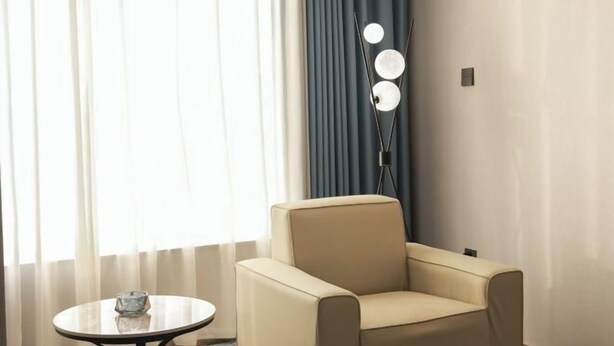Een kamer met een beige fauteuil, een kleine ronde tafel met een glazen object, en een vloerlamp met drie lampen.