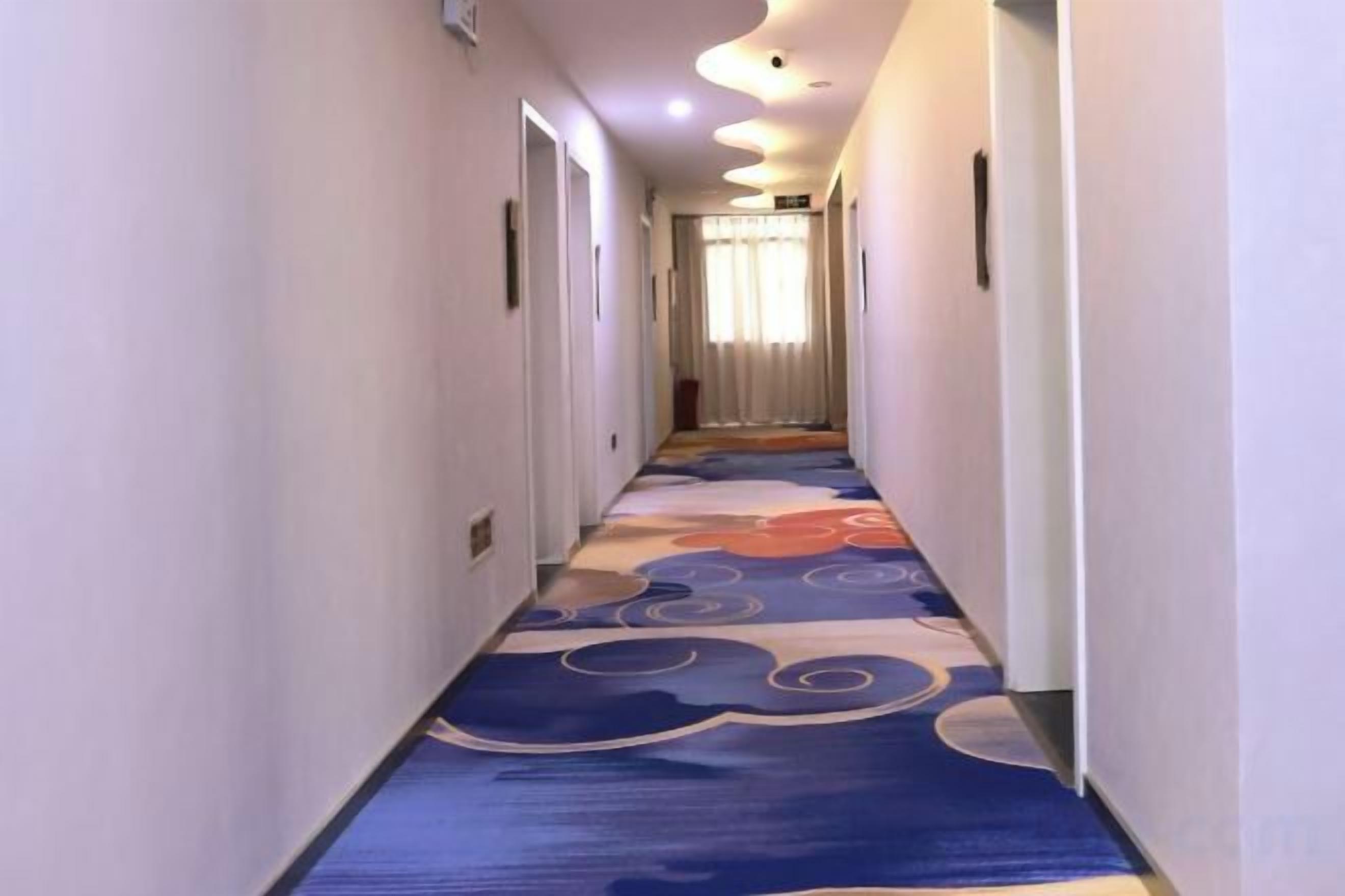 Un pasillo de hotel con alfombra estampada, puertas a ambos lados y una fuente de luz al final.